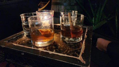A tray with four glasses: two filled with dark liquid, one with clear liquid, and one with a slice of orange.