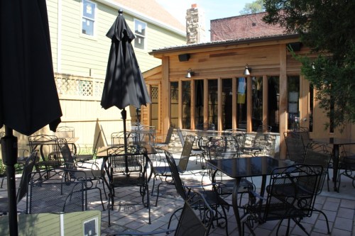 Outdoor seating area with black metal tables and chairs, shaded by umbrellas, next to a wooden building.