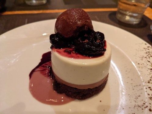 A layered dessert with chocolate and cream, topped with dark cherries and a scoop of chocolate sorbet on a white plate.
