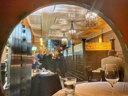 View through a circular vault door into a stylish restaurant with chandeliers and patrons dining.