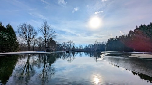 A serene lake reflects trees and a bright sun under a clear sky, with hints of ice along the shore.
