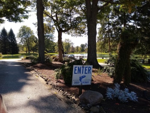 A sign reading "ENTER" at the entrance of a scenic park with trees and a pond in the background.
