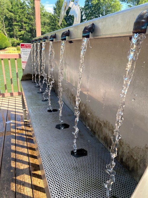 A metal water fountain with multiple spouts, each flowing water, set against a wooden deck and green trees in the background.
