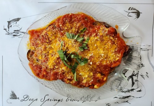 A glass dish with a colorful baked dish topped with tomato sauce, herbs, and cheese, on a decorative tablecloth.