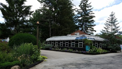 A greenhouse surrounded by lush greenery and trees, with a pathway leading up to it under a partly cloudy sky.
