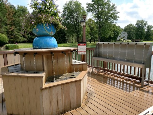 A decorative fountain with flowing water, surrounded by greenery and a bench, near a pond on a sunny day.