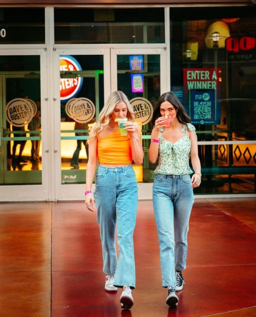 Two women walk towards the entrance of Dave & Buster's, each holding a drink and wearing casual outfits.
