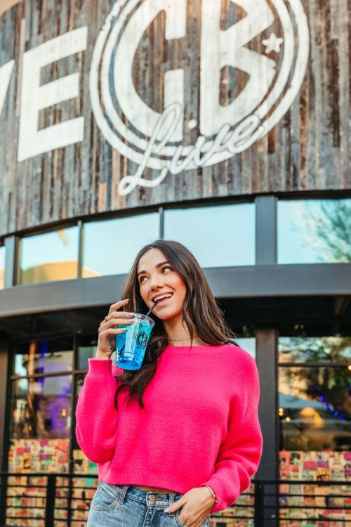 A woman in a pink sweater holds a drink, smiling in front of a rustic venue sign that reads "Live CB."