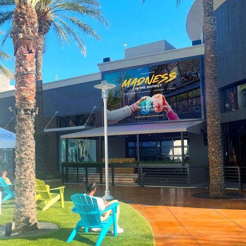 A vibrant outdoor scene featuring palm trees, colorful chairs, and a large advertisement for "Madness in the District."