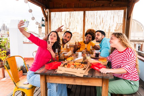 A group of five friends enjoying pizza and drinks on a rooftop, taking a selfie together.