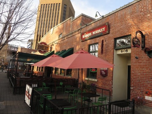 Exterior of Basque Market with outdoor seating and pink umbrellas, set against a brick building and city skyline.