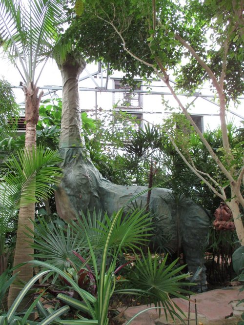 A large, green elephant sculpture surrounded by lush tropical plants in a greenhouse setting.