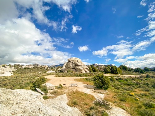 A scenic landscape featuring large rocks, green shrubs, and a blue sky with scattered clouds.