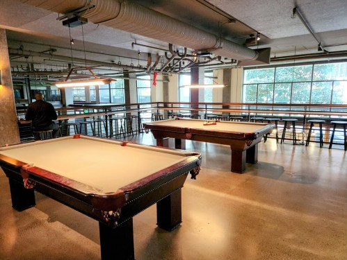 Two pool tables in a spacious, modern room with large windows and high-top seating in the background.