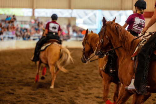 Three riders on horseback in a sandy arena, with a crowd watching in the background.