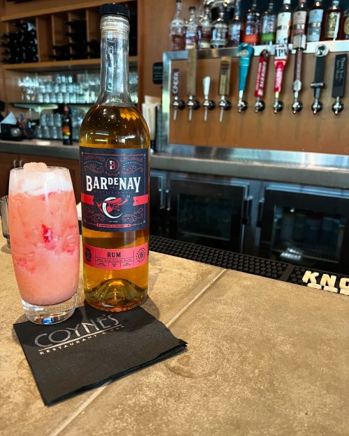 A bottle of Bardenay rum next to a pink cocktail in a glass on a bar counter, with a branded napkin.