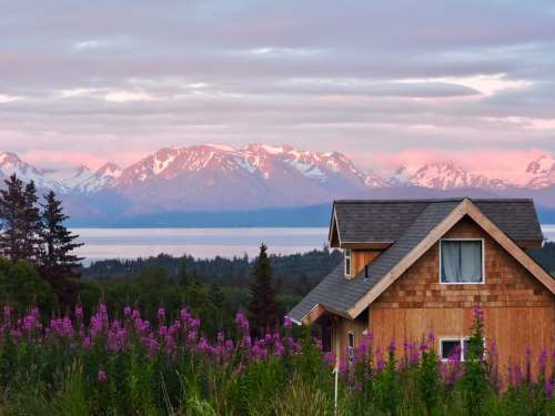 A cozy wooden house surrounded by purple flowers, with snow-capped mountains and a serene lake in the background at sunset.