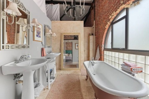 Spacious bathroom with a vintage bathtub, dual sinks, large mirror, and exposed brick walls. Natural light from windows.