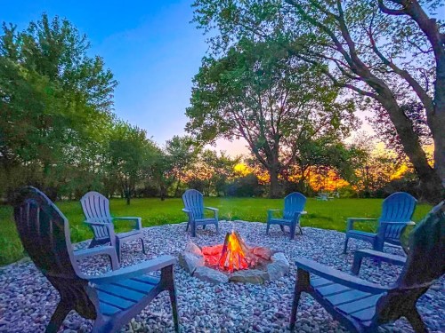 A cozy fire pit surrounded by blue chairs, set in a grassy area with trees and a colorful sunset in the background.