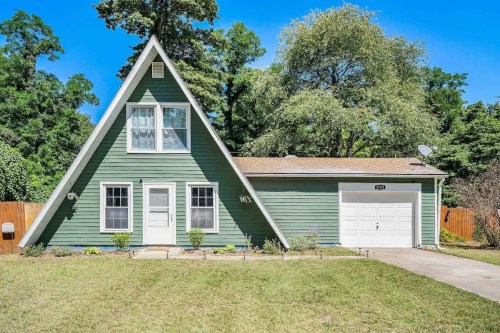 A green A-frame house with a garage, surrounded by trees and a grassy yard under a clear blue sky.
