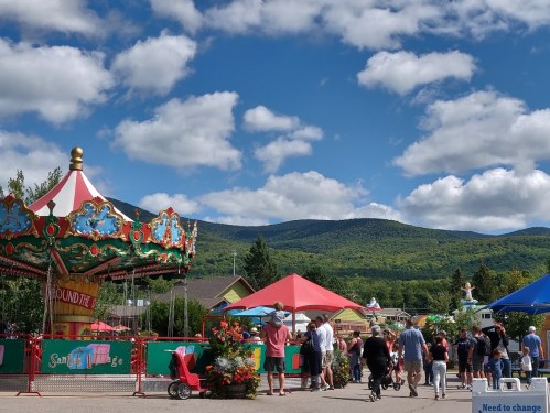 A vibrant carnival scene with a carousel, colorful tents, and people enjoying a sunny day against a mountain backdrop.