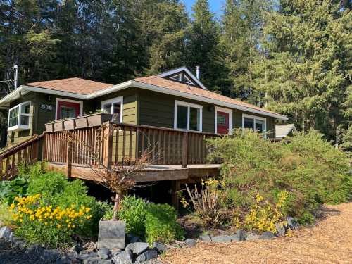 A green house with a wooden deck surrounded by trees and colorful flowers in a sunny outdoor setting.