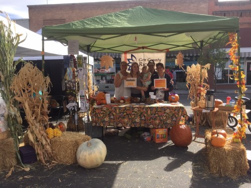 A festive market booth decorated with pumpkins and autumn decor, featuring three people and a baby.