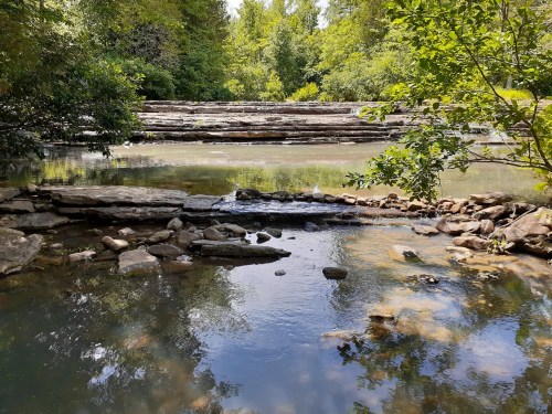 A serene river scene with smooth rocks, gentle water flow, and lush greenery reflecting in the calm water.