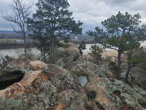 A rocky landscape with trees overlooking a river, under a cloudy sky.