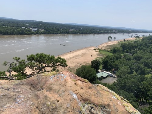 Aerial view of a riverbank with sandy shores, green trees, and distant hills under a clear blue sky.