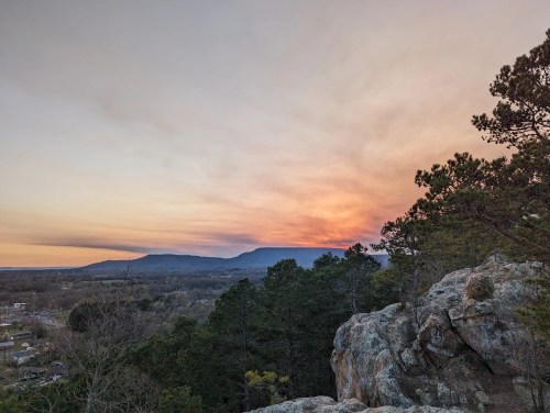 A scenic view of mountains at sunset, with colorful skies and trees in the foreground.