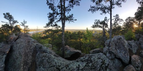 A panoramic view of a rocky landscape with trees, overlooking a river and distant hills under a clear sky.