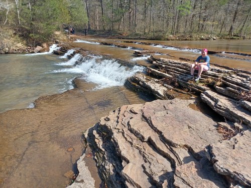 A person sits on rocky terrain by a flowing stream with small waterfalls, surrounded by trees in a natural setting.