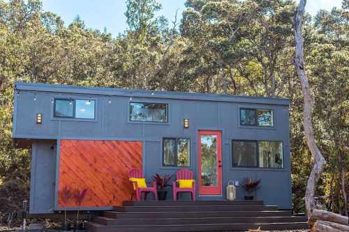 A modern tiny house with a wooden accent, bright red door, and colorful chairs on a deck, surrounded by trees.