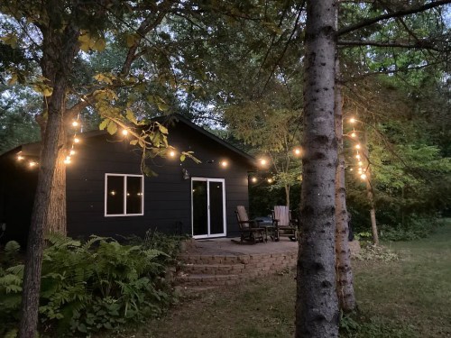 A cozy black cabin surrounded by trees, adorned with string lights, creating a warm evening atmosphere.