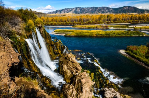 A scenic waterfall cascades over rocky cliffs into a river, surrounded by autumn foliage and mountains in the background.