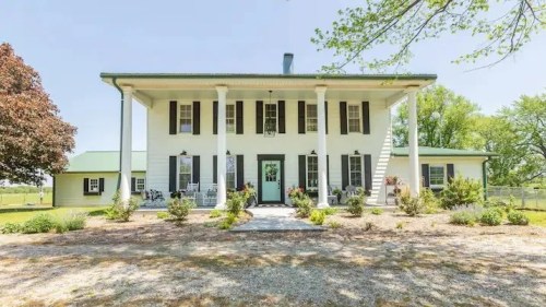 A large, two-story white house with green shutters, surrounded by greenery and a gravel pathway.