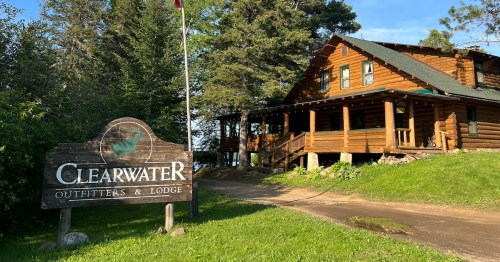 A rustic log cabin lodge with a sign reading "Clearwater Outfitters & Lodge" surrounded by trees and greenery.