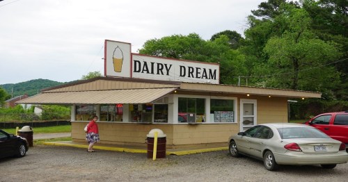 A small ice cream shop named "Dairy Dream" with a sign, surrounded by trees and parked cars. A person stands outside.