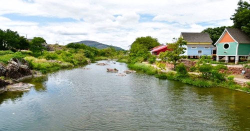 A serene river flows past colorful houses and lush greenery under a partly cloudy sky.