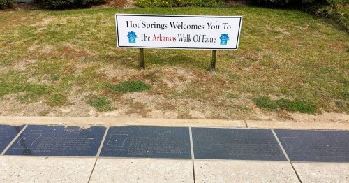 Sign welcoming visitors to Hot Springs and plaques for the Arkansas Walk of Fame on a grassy area.