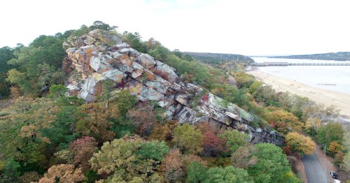Aerial view of a large rock formation surrounded by trees, with a beach and water in the background.