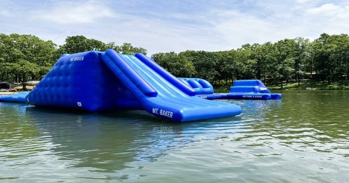 Two large blue inflatable water slides on a lake, surrounded by trees and a clear sky.