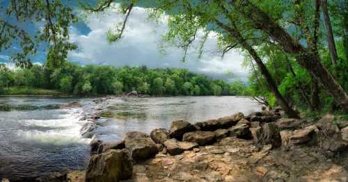 A serene river scene with lush green trees, rocky shore, and a cloudy sky reflecting on the water.