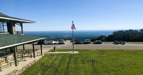 A scenic view from a building with a flag, overlooking a parking area and distant mountains under a clear blue sky.
