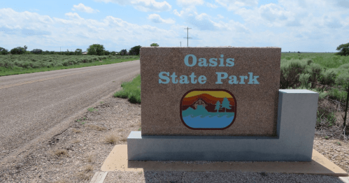 Sign for Oasis State Park beside a gravel road, with green fields and a blue sky in the background.
