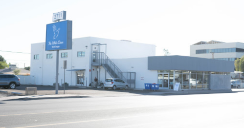 A modern building with a blue sign reading "The White Dove" and a parking area, located on a sunny street.