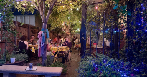 A cozy outdoor dining area with string lights, colorful tablecloths, and guests enjoying their meals under lush greenery.