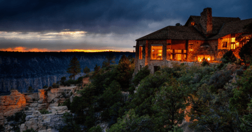 A rustic stone house on a cliff at sunset, surrounded by trees and dramatic clouds in the sky.