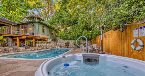 A serene backyard with a hot tub, swimming pool, and lush greenery, surrounded by a wooden fence and patio furniture.